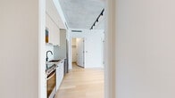This thoughtfully designed one-bedroom, one-bathroom residence delivers a modern urban aesthetic across a compact yet efficiently organized single floor. The open-concept kitchen and living area forms the heart of the unit, where light hardwood flooring, crisp white walls, and an exposed concrete ceiling create a striking industrial-contemporary contrast. Track lighting runs the length of the ceiling, providing adjustable illumination throughout. The kitchen is outfitted with light wood cabinetry, stainless steel appliances including a refrigerator, dishwasher, and oven range, along with a black matte faucet that adds a refined finishing touch. Floor-to-ceiling windows in the living area and bedroom flood the space with natural light and frame expansive city skyline views. The bedroom is a well-proportioned private retreat, complete with a dedicated closet featuring built-in shelving. The bathroom is finished in clean white tile with a glass-enclosed walk-in shower, black matte fixtures, and a modern vanity. An entrance hallway and additional hallway provide a smooth transition between rooms. The overall ambiance is clean, bright, and contemporary, making excellent use of every square foot.