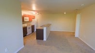 This single-floor space is designed with an open-concept layout that integrates the kitchen and living area into one cohesive, functional environment. The kitchen is equipped with white appliances, including a range, microwave, and refrigerator, all complemented by warm-toned wooden cabinetry that adds a classic touch. A dark-finished peninsula with a built-in breakfast bar serves as the natural dividing point between the kitchen and the living area, providing additional counter space and a casual dining option. Recessed lighting and a ceiling fan keep the space well-lit and comfortable throughout. The living area extends generously from the kitchen, featuring neutral-toned carpeting that adds warmth underfoot. Light-colored walls throughout create a clean, bright backdrop that enhances the sense of openness. The layout is highly practical, offering a smooth flow between functional zones, making the space well-suited for everyday living. The straightforward, unfussy design ensures that the interior is both welcoming and adaptable to a variety of furnishing styles.