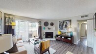 This single-floor residence offers a well-organized layout with distinct living, dining, kitchen, bedroom, and bathroom areas connected by a cohesive contemporary design. The living room serves as the central gathering space, anchored by a corner fireplace with a white mantel and flanked by a large sliding glass door that opens to a private balcony, allowing natural light to fill the room. Wood-style flooring runs throughout the main living areas, complementing the neutral gray walls and textured ceiling. A compact dining nook adjacent to the living space features hardwood-style flooring, pendant lighting, and wall décor, creating an intimate setting. The galley-style kitchen is efficiently arranged with dark espresso cabinetry lining both walls, light countertops, a subway tile backsplash, and black appliances including a range and refrigerator. Two bedrooms are finished with plush carpet, tufted headboard accommodations, closet access, and tasteful wall art. The bathroom includes a vanity with light countertops, a framed mirror, and a tub-shower combination. Throughout the residence, a consistent palette of gray, teal, and yellow accents ties each room together cohesively.