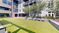 Located at 101 Washington St in Conshohocken, Pennsylvania, this thoughtfully designed outdoor amenity space offers a compelling blend of contemporary architecture and inviting landscape design. The courtyard is anchored by a vibrant, well-manicured synthetic lawn that provides an open and versatile gathering space, framed by raised planter beds filled with layered ornamental shrubs and native grasses that add color and texture throughout the seasons.

A standout feature of the space is the pergola-covered terrace, constructed with a bold dark steel framework and warm wood-toned deck tiles. Suspended swing chairs in a striking red finish serve as eye-catching focal points beneath the structure, offering a relaxed and unique seating experience. The terrace overlooks a lush, wooded ravine, providing sweeping natural views and a sense of privacy despite the urban setting.

Surrounding the lawn, additional seating areas are arranged along the perimeter, offering flexible spaces for socializing or quiet relaxation. Large glass-paneled doors connect the interior amenity spaces to the outdoors, ensuring a seamless indoor-outdoor flow. The multi-story residential building encloses the courtyard on multiple sides, with private balconies overlooking the shared grounds below. The overall ambiance is modern, serene, and meticulously maintained.