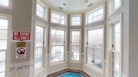 This single-floor space is defined by a standout indoor pool area that serves as its architectural centerpiece. The pool is encased in a sweeping bay window arrangement, featuring multiple double-hung windows framed in crisp white trim against soft gray walls. A row of transom windows above further amplifies the natural light, flooding the space with an airy, open ambiance. The surrounding tile work along the pool's edge adds a classic, refined touch, with warm earth tones complementing the neutral palette throughout. The walls carry a clean, light gray finish that lends a calm and welcoming atmosphere to the entire space. An entry area is accessible through a white panel door with brass hardware, accented by crown molding and a wall-mounted security light, suggesting a well-maintained and secure environment. The interior design favors traditional architectural elements — from the grid-patterned window panes to the paneled doors — creating a cohesive and timeless aesthetic. The combination of generous fenestration and thoughtful layout ensures the space remains bright and inviting throughout the day, with views of the surrounding outdoor landscape visible through the large windows.