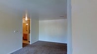 This single-floor space presents a straightforward and functional layout designed for comfortable everyday living. The main room is finished with wall-to-wall carpet in a neutral tone, providing a warm and quiet underfoot experience throughout. Smooth white walls and a clean white ceiling create a bright, open atmosphere, while a ceiling vent ensures consistent climate control. The room offers ample open floor space, making it highly adaptable to a variety of furniture arrangements and uses. A doorway visible from the main area leads directly into a bathroom, which features warm wood-toned cabinetry, a vanity with a mirror, and overhead lighting that lends a cozy, well-lit feel to the space. The transition between the main room and the bathroom is seamless, enhancing the overall flow of the layout. The neutral color palette throughout — from the carpeting to the walls — provides a versatile canvas that complements a wide range of interior styles. Overall, the space is clean, move-in ready, and well-suited for those seeking a practical and uncluttered environment.