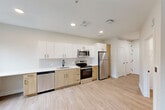 Located at 2111 Packard St in Ann Arbor, Michigan, this efficiently designed single-floor studio offers a thoughtful layout that maximizes its compact footprint. The open-plan living, dining, and kitchen area serves as the heart of the space, bathed in natural light from multiple windows that frame views of the surrounding brick buildings. Light hardwood-style flooring runs throughout, lending a clean, cohesive aesthetic.

The kitchen is well-appointed with a full suite of stainless steel appliances, including a refrigerator, electric range, microwave, and dishwasher. Cabinetry combines crisp white upper cabinets with warm natural wood lower cabinets, complemented by a white quartz countertop and a subway tile backsplash. Recessed lighting illuminates the space evenly overhead.

A hallway connects the main living area to the bathroom, closet, laundry, and utility spaces. The bathroom features a white vanity with a single sink, a large mirror with modern sconce lighting, black matte fixtures, and a walk-in shower with a black-framed glass door and tiled surround. A utility panel is discreetly integrated into the hallway wall. The dedicated laundry and closet areas add practical storage and convenience to this modern, move-in-ready space.