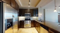 This single-floor residence offers a thoughtfully designed layout with high-quality finishes throughout. The kitchen is a standout feature, showcasing rich dark cabinetry with sleek hardware, granite countertops, a travertine tile floor, and a full suite of stainless steel appliances including a built-in refrigerator, range, microwave, and dishwasher. A pendant light fixture and recessed track lighting illuminate the space, which also features a tray ceiling and a tile backsplash for an elevated look. A kitchen island with a built-in wine rack adds both function and style, flowing seamlessly into the open living area.

The main living space is finished with warm hardwood-style flooring and large floor-to-ceiling windows that flood the room with natural light and offer sweeping views of palm trees and the surrounding cityscape. Track lighting enhances the bright, airy ambiance.

The bedroom features plush carpeting, a ceiling fan, and direct access to a private balcony with brick detailing and open city views. The primary bathroom is finished in travertine tile from floor to ceiling, and includes a soaking tub, a glass-enclosed walk-in shower, and a separate toilet area, creating a serene, spa-inspired retreat.
