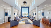 Located at 900 Lakeridge Rd in Hampton, Virginia, this single-floor community clubhouse delivers an impressive blend of elegance and functionality. The centerpiece of the space is a dramatic double-height great room anchored by a bold cobalt blue accent wall and floor-to-ceiling windows that flood the interior with natural light while offering views of the outdoor pool area. A striking contemporary chandelier hangs overhead, complementing the soaring ceilings and white decorative columns that frame the open layout. Rich, warm hardwood flooring flows throughout, tying the various areas together seamlessly. The main lounge is furnished with tufted upholstered seating, patterned armchairs, and a geometric area rug in blue, gold, and white, creating a welcoming social atmosphere. Adjacent to the lounge, a business center offers dual computer workstations set against navy and white striped drapery. A wood-front reception desk serves as the administrative hub of the space, positioned near a fully equipped kitchenette with stainless steel appliances, warm cabinetry, and pendant lighting. Private rooms accessible through glass-paned French doors provide additional flexible space. Large windows throughout ensure the entire clubhouse remains light-filled and airy from every vantage point.