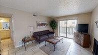 This single-floor residence offers a functional and comfortable layout with a cohesive, neutral aesthetic throughout. Light beige walls and large-format tile flooring unify the main living areas, creating a bright and easy-to-maintain foundation.

The living room serves as the central gathering space, featuring a tufted sofa, wrought iron and wood accent tables, and a colorful abstract canvas on the wall. A sliding glass door opens to a private balcony, allowing natural light to fill the room and providing a connection to the outdoors. A textured ceiling is present throughout the unit, and a ceiling fan overhead keeps the space comfortable.

Adjacent to the living room, the open dining area accommodates a round wooden table with iron chairs, positioned near a window with horizontal blinds that filter natural light. The layout flows seamlessly into the kitchen, which is defined by a raised bar counter with barstool seating — ideal for casual dining or entertaining. The kitchen is equipped with cabinetry and a full-size refrigerator.

The bedroom is carpeted for added warmth and comfort, with a sliding door leading to the balcony, offering outdoor access from multiple areas of the unit. A full bathroom features a tiled tub-and-shower combination, a toilet, built-in cabinetry, and clean, simple finishes throughout.