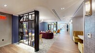 This thoughtfully designed single-floor amenity space offers a generous and well-appointed environment organized across distinct functional zones. The entrance greets visitors with wood-look tile flooring, a sleek glass-railed staircase, and a suspended linear pendant chandelier set against warm, textured walls — establishing an immediately polished first impression.

The main lounge area is a standout feature, anchored by a bold, curved burgundy sectional sofa atop a neutral area rug, surrounded by eclectic accent chairs in vibrant orange and earthy tones. A tall, black-framed open shelving unit serves as a stylish room divider, displaying books, plants, and decorative objects. Natural light filters in through large floor-to-ceiling windows, offering views of a landscaped outdoor courtyard and surrounding architecture.

A dedicated business center provides a long marble-topped workstation with multiple computer terminals, cognac leather task chairs, and warm pendant lighting — all oriented toward expansive windows that flood the room with daylight. A built-in lounge bench with colorful accent pillows and a striking abstract artwork complete this productive yet comfortable space.

An additional lounge nook features a built-in media wall with shelving, complemented by wingback chairs and soft carpeting. A separate dining area with a round dark table and blue velvet chairs connects seamlessly to adjacent spaces through glass-paneled doors, enhancing the overall flow and versatility of this amenity floor.