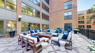 Located at 100 Paterson Plank Rd in Jersey City, NJ, this outdoor terrace courtyard is a thoughtfully designed communal space nestled alongside a striking multi-story brick building. The terrace features large-format stone tile paving throughout, providing a clean and durable foundation across the expansive open-air area.

Multiple seating arrangements are distributed across the space, including plush sofas and chairs outfitted with neutral cushions and vibrant blue accent pillows, centered around a substantial fire pit — ideal for gathering during cooler evenings. Additional seating clusters, including lounge chairs and a dining-style table, offer flexible use of the space for both relaxed lounging and casual outdoor dining.

Decorative planters filled with colorful flowers and lush greenery are positioned throughout, adding warmth and visual interest. A dark-railed perimeter fence defines the terrace boundary, beyond which mature trees provide a natural, verdant backdrop. Globe-style wall sconces mounted on the building facade contribute to ambient evening lighting.

The surrounding brick and steel-framed architecture, with expansive industrial-style windows, lends a distinctive urban character to the setting. The courtyard enjoys abundant natural light and open sky views, creating a welcoming outdoor retreat within an urban environment.