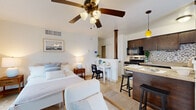 This single-floor studio is thoughtfully designed to maximize every square foot, offering a bright and cohesive open-plan layout that seamlessly connects the living, sleeping, and kitchen areas. The living space is filled with natural light, courtesy of a large sliding glass door that opens to an outdoor balcony and a secondary window framing outdoor views — both fitted with blinds and curtains for privacy. A ceiling fan with integrated lighting anchors the main living area, contributing to a warm, airy ambiance. Travertine tile flooring runs throughout, lending a clean, timeless aesthetic to the entire unit.

The kitchen is a standout feature, outfitted with rich dark-wood cabinetry, a mosaic tile backsplash, granite countertops, and stainless steel appliances including a range, microwave, and dishwasher. A peninsula with bar seating and a pendant light above serves as both a functional dining surface and a natural divider between the kitchen and living areas. A distinctive structural column adds architectural character to this transition space.

The bathroom maintains the same refined aesthetic, with matching travertine tile flooring, a dark-wood vanity topped with granite, a large mirror, and a full tub/shower combination. The overall ambiance of the space is calm, neutral, and well-appointed, making efficient use of its compact footprint.