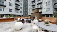 This expansive outdoor courtyard is nestled within a modern multi-story residential building, creating a sheltered yet open-air gathering space framed by the building's contemporary gray facade and glass-railed balconies. The ground level is finished with a striking large-format checkered stone tile pattern in contrasting shades of light and medium gray, lending a bold, geometric aesthetic underfoot.

A central outdoor fireplace, clad in textured brown stone, serves as a focal anchor for the courtyard's main lounge area, which features modular seating arranged over a colorful area rug. Nearby, round dining tables with white lattice chairs offer flexible seating for casual gatherings. Warm wood-paneled bench seating lines portions of the perimeter, adding a natural contrast to the otherwise contemporary material palette.

Sculptural oversized white globe and tall tapered planters are distributed throughout the space, introducing greenery and organic forms that soften the hardscape. A wicker dome pod chair and bright blue sculptural lounge seating add playful design accents to one corner. A BBQ and outdoor kitchen station with a wooden surround provides functional amenity space. The courtyard benefits from open sky above, offering natural daylight throughout the day.