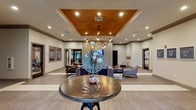 Located at 6721 Washington Ave in Ocean Springs, Mississippi, this single-floor clubhouse facility offers a thoughtfully designed communal space that combines modern elegance with comfortable functionality. Upon entry, a grand foyer greets visitors with large-format tile flooring, recessed lighting, and a striking round accent table beneath a coffered ceiling detailed with a warm copper finish — an immediately eye-catching architectural element that sets the tone for the entire space.

The open-concept lounge area flows naturally from the entry, featuring wood-look plank flooring, a boldly patterned area rug, and a curated arrangement of seating including a plush cream sofa, leather accent chairs, and glass and metal coffee tables. A rope-wrapped geometric chandelier with Edison-style bulbs anchors the lounge visually, casting a warm, inviting glow. A wall-mounted flat-screen television is framed by textured tile accent wall panels, creating a polished focal point for gathering and relaxation.

Additional seating areas extend throughout the space, offering varied arrangements suited for both social interaction and quiet retreat. The fitness center, visible through a glass-paneled door, provides convenient access to exercise equipment. Neutral wall tones, dark wood trim accents, crown molding, and consistent recessed lighting unify the design, creating a cohesive, upscale ambiance throughout this well-appointed community space.