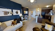 This single-floor residence showcases a thoughtfully designed open-concept layout that seamlessly connects the living, dining, and kitchen areas into one cohesive and functional space. The living area is anchored by a striking deep navy accent wall, which adds a bold, contemporary contrast to the neutral carpeted flooring and lighter surrounding walls. A ceiling fan with a modern light fixture keeps the space comfortable, while carefully selected furnishings — including a plush sofa, leather lounge chair, and accent tables — contribute to a warm yet sophisticated ambiance. The dining area transitions naturally from the living space, offering a casual setting ideal for everyday meals or entertaining guests. The kitchen stands out with its granite countertops, dark cabinetry, and a peninsula breakfast bar fitted with bar-height seating, providing both additional prep space and a social gathering point. Pendant lighting above the bar adds a refined finishing touch. A glimpse beyond the main living area reveals a hallway leading to additional rooms, suggesting a well-organized and practical floor plan. Throughout the space, the interplay of rich, dark tones against bright whites and neutral carpeting creates an inviting atmosphere that balances modern style with everyday livability.