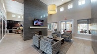 This contemporary communal space is designed to impress, featuring dramatic soaring ceilings that amplify the sense of openness throughout. The centerpiece of the main lounge is a striking floor-to-ceiling tiled fireplace wall clad in large-format dark grey tile, paired with a wall-mounted flat-screen television — creating an anchor for the primary seating area. Patterned armchairs arranged around a low coffee table invite relaxed conversation, all set atop an area rug over light-toned tile flooring that runs throughout the space.

Large windows positioned high along the walls flood the interior with abundant natural light, while oversized drum pendant lights and exposed Edison-bulb pendants provide warm ambient illumination. A secondary lounge area offers comfortable grey sofas accented with vibrant orange throw pillows, complemented by colorful abstract artwork adorning the walls. High-top bar tables and stools line the perimeter, providing flexible seating suited for casual work or social gatherings.

A dedicated beverage station features sleek dark cabinetry, a stone mosaic tile backsplash, light quartz countertops, and a built-in mini-refrigerator, offering a convenient self-serve coffee and refreshment setup. Glass-paneled doors connect the interior to exterior areas, enhancing the flow between indoor and outdoor environments. The overall aesthetic is polished, modern, and welcoming.