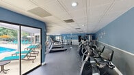 Located at Royal Park in Carrboro, North Carolina, this fitness center is a thoughtfully designed exercise space that provides residents with a comprehensive workout facility. The room is finished with a distinctive coffered ceiling and two-tone blue walls accented by the Royal Park fleur-de-lis branding, creating a clean, modern aesthetic throughout.

The space is generously equipped with a wide array of fitness equipment to accommodate varied workout routines. A row of treadmills and elliptical machines lines one side of the room, ideal for cardiovascular training, while stationary bikes add further cardio options. The strength training area features a cable machine, a squat rack, a dumbbell rack with a full set of free weights, and a weight bench, supporting a complete resistance workout.

Durable rubber flooring covers the main exercise area, transitioning to hardwood in a secondary section that houses additional free weights, medicine balls, and a stability ball — well-suited for functional training and stretching. Full-length mirrors along one wall enhance the sense of space and allow for proper form checks during workouts.

Large sliding glass doors and expansive windows flood the interior with natural light and offer direct views of the outdoor swimming pool and surrounding green landscaping, creating an inviting and energizing atmosphere. A wall-mounted television and water station are also present for added convenience.