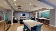 Located in the heart of Cincinnati at 215 Ninth St E, this single-floor amenity space offers a well-appointed and cohesive environment designed for both relaxation and entertainment. The open layout flows seamlessly between several distinct zones, all unified by warm wood-look flooring, a neutral gray color palette, and recessed drop-panel ceilings with abundant overhead lighting.

The centerpiece of the main lounge area is a striking white stacked-stone accent wall, which frames both a wall-mounted flat-screen television and a sleek linear electric fireplace below it. Comfortable seating, including a leather sofa and accent chair, creates an inviting gathering spot. Adjacent to the lounge is a dining area furnished with a large rectangular table and teal upholstered chairs, adding a vibrant pop of color throughout the space.

A wet bar area features white countertops, bar-height seating, and a stainless steel refrigerator, accented by a bold teal wall that adds visual interest. Pendant lighting overhead enhances the social atmosphere of this serving station.

A separate billiards room offers a full-size pool table beneath a decorative hanging light fixture, with natural light filtering in through a large window with views of the surrounding urban landscape. Throughout the space, curated artwork and vintage-style posters contribute to the lively, contemporary character of this exceptional amenity.