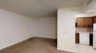 This single-floor space offers a clean and functional layout designed for straightforward living. The main room is finished with wall-to-wall carpet in a warm neutral tone, providing a soft underfoot feel throughout. The walls are painted in a crisp, bright white, contributing to a simple and uncluttered atmosphere that allows for flexible furnishing arrangements. A textured ceiling adds a subtle decorative detail overhead. A solid white entry door is positioned along one wall, providing direct access to the space and featuring a deadbolt lock for added security. White baseboards run along the perimeter of the room, offering a clean transition between the walls and carpeted floor. Electrical outlets and light switch plates are positioned at standard heights along the walls, ensuring practical usability throughout. The overall ambiance of the space is understated and neutral, creating a blank canvas well-suited for personalization. The open layout allows for versatile use of the available area, whether for a living space, sleeping area, or a combination of functions. The minimalist design keeps the focus on utility and adaptability.
