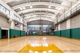 This expansive indoor basketball court is a well-appointed athletic facility designed for serious play and versatile use. The court features a regulation-sized hardwood maple floor with clearly marked playing lines in white and black, complemented by bold yellow paint in the key areas, creating a visually striking contrast against the natural wood finish.

The architecture is defined by a dramatic vaulted ceiling supported by a white structural framework, lending the space a generous, open feel with considerable vertical clearance. Suspended basketball hoops with glass backboards are installed on both ends and along the sides, accommodating multiple court configurations. Overhead, globe pendant lighting and additional ceiling-mounted fixtures ensure consistent, even illumination throughout.

The perimeter walls are lined with thick green padded panels for player safety, topped by neutral-toned acoustic wall tiles that contribute to sound management within the space. A large floor-to-ceiling glass curtain wall on one end opens the court visually to an adjacent atrium filled with natural light and greenery. A steel staircase leads to an upper mezzanine level overlooking the court. Additional amenities include a digital scoreboard, equipment storage rack, and a roll-up utility door for convenient access.