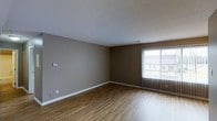 This single-floor space offers a clean, straightforward layout designed for comfortable everyday living. The main living area immediately draws attention with its warm hardwood-style flooring, which adds a sense of warmth and continuity throughout the room. Neutral, earthy-toned walls in a soft taupe complement the flooring and create a calm, understated ambiance that serves as a versatile backdrop for a variety of décor styles. A large window fitted with vertical blinds allows ample natural light to fill the space, brightening the interior and offering a view of the surroundings outside. The textured ceiling adds a subtle architectural detail overhead, while crisp white baseboards provide a clean contrast against the wall color. A wall-mounted thermostat is conveniently positioned, indicating climate control for year-round comfort. The open, uncluttered layout provides flexibility in how the space can be arranged and utilized. Overall, this space presents a practical and inviting environment, with its combination of natural light, durable flooring, and neutral finishes making it well-suited for a range of uses.