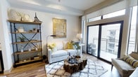 This single-floor space showcases a thoughtfully designed living area that blends modern and rustic aesthetics seamlessly. The room is anchored by a light-toned sofa dressed with vibrant yellow and botanical-patterned accent pillows, paired with a striking natural wood stump coffee table that adds an organic, earthy contrast to the otherwise contemporary setting. A plush geometric-patterned area rug grounds the seating arrangement, adding texture and warmth to the wood-look flooring beneath.

An industrial-style open shelving unit lines one wall, displaying curated decorative objects and personal items, contributing to the space's layered character. Above the sofa, a large abstract canvas in gold and neutral tones serves as a bold focal point. A floor lamp and a side-table lamp provide warm, ambient lighting throughout.

Natural light floods the room through a full-height sliding glass door that opens onto a private balcony, offering outdoor seating and views beyond. Neutral walls and clean architectural lines keep the space feeling bright and open, while the carefully selected furnishings and décor create an inviting, well-curated atmosphere.