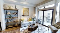 This single-floor space showcases a thoughtfully designed living area that blends modern and rustic aesthetics seamlessly. The room is anchored by a light-toned sofa dressed with vibrant yellow and botanical-patterned accent pillows, paired with a striking natural wood stump coffee table that adds an organic, earthy contrast to the otherwise contemporary setting. A plush geometric-patterned area rug grounds the seating arrangement, adding texture and warmth to the wood-look flooring beneath.

An industrial-style open shelving unit lines one wall, displaying curated decorative objects and personal items, contributing to the space's layered character. Above the sofa, a large abstract canvas in gold and neutral tones serves as a bold focal point. A floor lamp and a side-table lamp provide warm, ambient lighting throughout.

Natural light floods the room through a full-height sliding glass door that opens onto a private balcony, offering outdoor seating and views beyond. Neutral walls and clean architectural lines keep the space feeling bright and open, while the carefully selected furnishings and décor create an inviting, well-curated atmosphere.