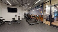 This fitness center is designed to deliver a comprehensive workout experience within a thoughtfully organized single-level space. The layout is divided into distinct functional zones, each outfitted with professional-grade equipment to accommodate a wide range of training styles.

The cardio area features a row of Matrix treadmills, stationary cycling bikes, and elliptical machines lined against a dramatic large-scale mural depicting a runner in a desert landscape, creating an immersive and motivating environment. A wall-mounted flat-screen television and a hydration station with a water fountain are conveniently positioned nearby.

The strength and functional training zone is anchored by a multi-purpose cable and functional trainer rack stocked with TRX suspension systems, kettlebells, medicine balls, and a full dumbbell rack. A large mirrored wall expands the sense of space and allows users to monitor their form. An adjustable weight bench completes the setup.

A striking snowboarder mural dominates one wall of the free-weight area, adding an energetic visual focal point. Overhead, large industrial ceiling fans and linear LED lighting ensure the space remains well-ventilated and brightly lit. Durable rubber flooring throughout provides traction and impact absorption, reflecting the facility's practical, performance-focused design.