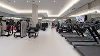 This spacious fitness center is designed with a clean, modern aesthetic that creates an inviting and professional environment for exercise. The layout is thoughtfully organized into distinct zones, separating cardio equipment from strength-training machines to allow for efficient movement throughout the space. The tray ceilings, finished in a neutral gray tone, incorporate recessed lighting alongside warm ambient cove lighting that runs along the perimeter, contributing to a polished and well-lit atmosphere.

The cardio zone features a full row of treadmills, elliptical machines, and stationary bikes—all equipped with integrated display screens—positioned alongside a floor-to-ceiling mirror wall that enhances the sense of space. Wall-mounted televisions provide entertainment options for users. The strength training area is well-stocked with a variety of cable machines, resistance equipment, adjustable benches, and functional training stations, including TRX suspension systems.

The flooring throughout is a light-colored, smooth surface that is easy to maintain and visually expands the room. Patterned wallcovering accents and structural columns add subtle architectural character. The overall ambiance is clean, upscale, and conducive to focused, effective workouts.