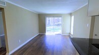 This single-floor space offers a clean, functional layout with a welcoming sense of openness. The living area is anchored by rich, dark hardwood flooring that adds warmth and character throughout. Crown molding runs along the ceiling perimeter, lending a classic, polished finish to the space.

Natural light is a defining feature, with a large sliding glass door fitted with vertical blinds serving as a focal point along one wall. This door opens to a view of a verdant outdoor area, seamlessly connecting the interior with the greenery outside. An additional window on the adjacent wall further enhances the brightness and airiness of the room.

The walls are finished in a soft, neutral tan tone that complements the warm flooring and white trim work, creating a cohesive and inviting palette. The textured ceiling and crisp white baseboards complete the interior detailing. The overall ambiance is light, airy, and well-maintained, making the space versatile and move-in ready. The layout provides ample room for furnishing and personalization, while the natural light and outdoor connection add a refreshing, livable quality to the environment.