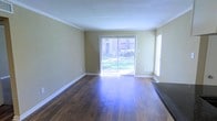 This single-floor space offers a clean, functional layout with a welcoming sense of openness. The living area is anchored by rich, dark hardwood flooring that adds warmth and character throughout. Crown molding runs along the ceiling perimeter, lending a classic, polished finish to the space.

Natural light is a defining feature, with a large sliding glass door fitted with vertical blinds serving as a focal point along one wall. This door opens to a view of a verdant outdoor area, seamlessly connecting the interior with the greenery outside. An additional window on the adjacent wall further enhances the brightness and airiness of the room.

The walls are finished in a soft, neutral tan tone that complements the warm flooring and white trim work, creating a cohesive and inviting palette. The textured ceiling and crisp white baseboards complete the interior detailing. The overall ambiance is light, airy, and well-maintained, making the space versatile and move-in ready. The layout provides ample room for furnishing and personalization, while the natural light and outdoor connection add a refreshing, livable quality to the environment.