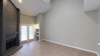 This single-floor space is defined by its vaulted ceilings, which lend a sense of openness and grandeur to the main living area. The walls are finished in a neutral greige tone, creating a calm, versatile backdrop suited to a variety of interior styles. Light-toned hardwood-style flooring runs throughout, adding warmth and visual continuity to the space.

A standout architectural detail is the built-in shelving unit recessed into the wall, providing both functional storage and a decorative focal point without occupying additional floor space. Adjacent to the shelving, French doors with white shuttered blinds allow natural light to filter into the room while offering privacy and the option to connect with the outdoor area beyond.

The vaulted ceiling rises to an impressive height along one wall, accentuating the spacious feel of the interior. Clean white baseboards contrast neatly against the neutral walls and flooring, contributing to a polished, well-maintained aesthetic. Overall, the space presents an open, bright, and functional layout with thoughtful architectural features that enhance both its practicality and visual appeal.