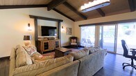 This single-floor space showcases a warm, rustic aesthetic anchored by a striking vaulted ceiling with dark-stained exposed wood beams and natural wood paneling, creating an inviting, lodge-inspired ambiance. The living area is generously sized, comfortably accommodating a large sectional sofa and a sturdy wooden coffee chest that doubles as a centerpiece and storage piece. A wall-mounted flat-screen television sits atop a solid wood media console, flanked by wall sconces that add a soft, ambient glow. A woven accent chair with striped cushions provides additional seating near a decorative wooden artwork piece, adding to the layered, nature-inspired decor. Recessed lighting and a pendant chandelier complement the natural tones throughout. Sliding glass doors along one wall flood the space with natural light while offering a direct view of and access to an outdoor patio area, where lounge seating is visible. The seamless transition between the interior and exterior enhances the overall sense of openness. Dark hardwood flooring grounds the space, tying together the warm earth tones and natural textures present throughout the room.