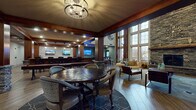Located at 9815 Seasons West Dr. in Indianapolis, Indiana, this single-floor community clubhouse blends rustic craftsmanship with upscale design. The grand entrance foyer sets an immediate tone of sophistication, featuring herringbone-patterned hardwood flooring, decorative wainscoting, rich dark wood columns with stacked stone bases, and a sweeping staircase with wrought iron balusters beneath soaring coffered ceilings.

The main gathering area is anchored by a dramatic floor-to-ceiling stacked stone fireplace flanked by oversized multi-pane windows that draw in abundant natural light and offer views of the surrounding landscape. Seating areas are thoughtfully arranged throughout, including a curved lounge sofa accented with gold sculptural wall art, leather arm chairs, and a substantial round dining table beneath an ornate chandelier.

A media lounge area features multiple wall-mounted flat-screen TVs with rows of seating, ideal for group viewing. An open kitchen includes dark granite countertops, warm wood cabinetry, a stone tile backsplash, stainless steel appliances, and stylish pendant lighting. A billiards table and decorative open shelving add recreational and aesthetic character to the space. The clubhouse presents a warm, welcoming ambiance suited for both social gatherings and quiet relaxation.
