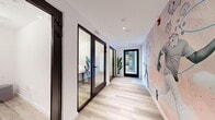 Located at 100 Hale Ave in White Plains, NY, this contemporary office suite offers a thoughtfully designed single-floor layout comprising four rooms. A central corridor serves as the spine of the space, finished with light wood-toned flooring and crisp white walls that create a clean, professional atmosphere. One wall of the hallway is adorned with a striking artistic mural — a whimsical, hand-illustrated design in soft blush and neutral tones — adding a creative energy to the space.

The rooms are enclosed by sleek, floor-to-ceiling glass partitions with bold black metal frames, allowing natural light to flow freely throughout while maintaining acoustic separation. Each room is accessible via full-height glass doors, lending an open and transparent feel to the overall layout.

One of the rooms functions as a conference or meeting space, furnished with a wooden table, seating for several occupants, a wall-mounted display screen, and large windows overlooking the surrounding neighborhood — providing ample natural light and an exterior view. Another area features a vibrant mural accent wall with a surrealist design, complemented by lush tropical plants that introduce a lively, biophilic element.

Recessed ceiling lighting and clean architectural lines throughout reinforce the modern, professional character of this well-appointed office suite.