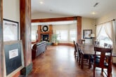 This well-appointed single-floor space offers a generous, open layout that seamlessly connects the living room, dining area, and kitchen into one cohesive environment. The interior is defined by its striking architectural character, most notably the bold, richly stained wood columns and ceiling beams that frame the open-plan living area and lend a rustic yet refined warmth throughout.

The living room features vaulted ceilings, a corner stone fireplace with a substantial wood mantel, and a sweeping bank of windows that floods the space with natural light and provides broad exterior views. The polished, stained concrete floors in deep amber and mahogany tones extend throughout, grounding the space with a durable and visually rich surface.

The dining area sits adjacent to the living room, benefiting from large windows dressed with sheer linen curtains that soften the natural light. A substantial wood dining table anchors this zone, making it well-suited for gatherings.

The kitchen is efficiently designed, featuring warm knotty wood cabinetry, dark countertops, stainless steel appliances, and a window above the sink that brings in additional daylight. The overall ambiance of the space is inviting and character-rich, blending natural materials, abundant light, and an open, connected floor plan into a highly functional and visually cohesive environment.