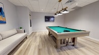 Located at 507 E Main St in Bound Brook, NJ, this thoughtfully designed single-floor space serves as a polished recreational retreat. The interior is unified by light-toned luxury vinyl plank flooring and soft gray walls, creating a clean, contemporary aesthetic throughout.

At the heart of the space is a full-size billiard table with a teal felt surface and a rustic wood frame, illuminated by a striking brass and black pendant light fixture. A wall-mounted flat-screen TV is positioned nearby, complementing the entertainment-focused atmosphere. A cue rack mounted on the wall keeps equipment organized and accessible.

A comfortable lounge area offers a streamlined neutral sofa accented by a bold abstract painting in blue and gold tones, along with a round glass-top side table — providing an inviting spot to relax between games.

A built-in wet bar area features white shaker-style cabinetry, a white countertop, a stainless steel mini refrigerator, and a sink, all highlighted by sleek black pendant lighting and a round mirror mounted above. A bar-height chair completes this functional serving station.

The space also includes a small dining or work area with a white table and chairs. The drop ceiling with recessed lighting and integrated ventilation ensures a well-lit, comfortable environment throughout.