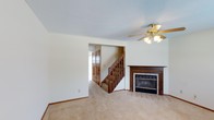 This two-story home offers a well-structured layout designed for comfortable everyday living. The main living area is finished with wall-to-wall carpeting that lends a warm, inviting feel to the space. A standout feature of this room is the classic wood-mantled fireplace, framed by tile detailing, which serves as a natural focal point and provides a cozy atmosphere. A ceiling fan with a light fixture is centrally positioned overhead, contributing to both comfort and ambient lighting. The textured ceiling adds a traditional character throughout. A wooden staircase with a classic balustrade connects the main level to the upper floor, with a view into an adjacent hallway featuring built-in storage. The transition between rooms flows naturally, giving the home a cohesive and functional feel. The neutral color palette of warm whites and beiges throughout the main level creates a clean, versatile backdrop well-suited to a variety of interior styles. The two-floor layout provides clear separation between living and private spaces, making this home practical and adaptable for a range of needs.