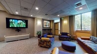 Located at 12 Park Pl in Hartford, Connecticut, this single-floor space is designed to serve as a functional and comfortable communal gathering area. The layout is divided into two primary zones: a spacious lounge area and an adjoining kitchenette.

The lounge area is generously appointed with an eclectic mix of seating, including multi-patterned armchairs, a long upholstered sofa with accent pillows, and a rounded ottoman, all arranged to encourage conversation and relaxation. A large wall-mounted flat-screen television serves as a central focal point, complemented by a simple upholstered bench beneath it. Colorful framed artwork adorns the walls, adding visual interest and warmth to the space. Potted indoor plants are thoughtfully placed throughout, bringing a touch of greenery to the interior. Drape-framed windows allow natural light to filter in, offering views of the surrounding greenery outside. Recessed lighting and wall sconces provide warm, layered illumination throughout.

The kitchenette features sleek dark cabinetry lining the walls, housing a microwave, sink, and full-size refrigerator. A curved high-top counter with bar stools provides an additional casual dining or gathering option, while framed artwork and a decorative floral centerpiece enhance the area's welcoming atmosphere. Light tile flooring contrasts cleanly with the lounge's carpeted areas, clearly delineating the two functional zones.