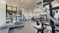 This single-floor fitness center is thoughtfully designed to provide a comprehensive workout environment. The space is anchored by a striking geometric mural in teal, blue, and purple tones that spans an entire accent wall, lending a dynamic and energetic ambiance to the room. Durable dark rubberized flooring covers the workout area, providing both safety and functionality throughout.

The cardio zone features a treadmill, elliptical trainer, and a stationary recumbent bike, offering a well-rounded selection for aerobic exercise. The strength training area is equally well-appointed, with multiple cable and weight machines, adjustable benches, and a multi-function cable tower to support a full range of resistance workouts.

A full wall of floor-to-ceiling mirrors expands the sense of space and allows users to monitor their form during exercise. Clerestory windows along one wall invite natural light, while recessed ceiling lighting ensures consistent illumination throughout. A ceiling fan aids in air circulation across the main workout floor.

A distinct glass-partitioned section with textured panels creates a semi-private zone within the facility, housing additional equipment. The clean white walls and open layout contribute to an uncluttered, professional atmosphere well-suited for focused training sessions.