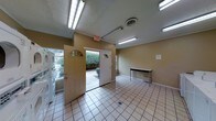 This communal laundry facility is designed with functionality and convenience in mind. The space features light beige tile flooring throughout, providing a clean and easy-to-maintain surface. Bright fluorescent lighting is mounted across the ceiling, ensuring the area is well-illuminated at all times. The textured ceiling adds a modest architectural detail, while crown molding along the upper walls lends a subtle finishing touch.

Two white entry doors with arched transom windows allow natural light to filter in, creating a welcoming atmosphere while maintaining clear access points for residents. A designated folding table is positioned along one wall, offering a convenient surface for sorting and folding laundry. Laundry machines are accessible along the perimeter of the space, and a utility drain is centrally located in the floor for easy maintenance.

The walls are painted in a warm neutral tone, contributing to the overall clean and orderly feel of the room. An illuminated exit sign and a visible HVAC vent reflect the facility's attention to safety and climate comfort. The layout is open and spacious, allowing multiple users to move freely and comfortably. Overall, this laundry facility is a practical and well-maintained shared amenity.