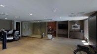 This single-floor space showcases a modern, open-plan design that seamlessly integrates multiple functional zones within one expansive room. The most striking feature is a large, floor-to-ceiling glass-enclosed shower or wet room, which occupies a prominent portion of the space. Its fully transparent glass walls allow for an unobstructed view across the room, contributing to an airy, spa-like atmosphere. The enclosure includes plumbing fixtures and appears to accommodate generous movement.

Adjacent to the glass enclosure, a warm wood-paneled built-in shelving unit provides display and storage space, complemented by a wall-mounted flat-screen television that anchors the entertainment area. To the right, a compact wet bar or utility sink station with cabinetry adds further functionality to the space. Decorative artwork adds a personal touch to the neutral-toned walls.

The flooring throughout is light-toned hardwood, enhancing the sense of openness and warmth. Recessed ceiling lighting is distributed evenly, providing consistent, well-distributed illumination across the entire area. The overall ambiance is clean, contemporary, and thoughtfully designed.
