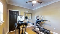 This single-floor fitness space is thoughtfully designed to deliver a functional workout environment. The room features hardwood flooring partially covered by an exercise mat, providing both durability and comfort during workouts. Two elliptical cross-trainer machines are positioned as the centerpiece of the space, each equipped with digital display consoles for tracking workout metrics. A large wall-mounted mirror enhances the sense of space and allows users to monitor their form during exercise. The room is finished with elegant crown molding and warm, neutral-toned walls that contribute to an inviting atmosphere. Overhead, a ceiling fan with an integrated light fixture ensures adequate air circulation and lighting during workouts. A solid dark-paneled door provides a private, enclosed environment. The overall layout is efficient and well-organized, making the most of the available square footage. This fitness room is well-suited for residential or small commercial use, offering a convenient and comfortable setting for daily cardiovascular exercise without the need to leave the building.