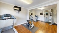 Located at 872 Bettina Ct in Houston, TX, this fitness center is a well-equipped, single-floor exercise space designed to support a variety of workout routines. The room features warm hardwood flooring throughout, complemented by clean white walls adorned with crown molding that lend a polished, finished look to the space.

The fitness center is divided into two connected areas. The main zone houses cardio equipment, including treadmills and an elliptical machine, positioned to take advantage of natural light streaming in through windows fitted with blinds. A wall-mounted flat-screen television provides entertainment during workouts. A wall-mounted fan ensures comfortable air circulation within the space.

An adjoining alcove, framed by a wide doorway, extends the workout area and features stationary bikes, a multi-function cable resistance machine with various attachments, and a large floor-to-ceiling mirror that visually expands the room and allows users to monitor their form. Protective rubber floor mats are placed beneath the equipment throughout. Posted fitness center rules and informational signage reflect the space's shared-use nature.

Flush-mounted ceiling light fixtures provide consistent overhead illumination, while a glass-paneled exterior door allows additional natural light and outdoor views of the surrounding greenery, creating a bright and motivating environment for exercise.