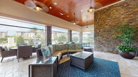 Located at 906 Oasis Palm Cir in Ocoee, Florida, this single-level covered outdoor lounge area is a well-appointed communal gathering space designed for relaxed resort-style living. The space is anchored by a striking stacked-stone accent wall that houses a built-in fireplace topped with a wall-mounted flat-screen television, creating a compelling focal point for the seating area. Rich wood-plank ceilings extend throughout, fitted with recessed lighting and pendant fixtures that lend a warm, inviting ambiance. Travertine tile flooring adds a natural, upscale aesthetic that flows seamlessly across the entire space. The lounge is furnished with wicker sectional seating and accent chairs arranged over a teal geometric area rug, complemented by glass-top wicker coffee tables. Large open-air window openings and floor-to-ceiling glass panels flood the interior with abundant natural light while providing unobstructed views of the outdoor pool area, palm trees, and manicured grounds beyond. Additional amenities include a ping-pong table, lush potted tropical plants that enhance the Florida atmosphere, and multiple separate seating arrangements to accommodate various group sizes. The overall design blends natural materials with modern comfort, resulting in an airy and functional outdoor living space.