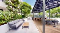 Located at 16 N Federal Hwy in Dania Beach, Florida, this outdoor amenity level showcases a thoughtfully designed resort-style courtyard framed by a modern, multi-story white building with floor-to-ceiling windows and private balconies. At its heart lies a sparkling pool flanked by manicured green turf and a pair of slatted wood-and-metal cabanas, providing shaded relaxation areas ideal for enjoying the South Florida sunshine.

The surrounding courtyard is richly landscaped with mature palm trees, tropical canopy trees, and lush planters brimming with vibrant greenery, creating a sense of privacy and seclusion. Large-format porcelain pavers connect the various outdoor zones seamlessly, lending a clean, upscale aesthetic throughout.

A covered pergola area features a decorative tiled outdoor kitchen and grilling station, complemented by a mix of rope-woven lounge seating, teak coffee tables, and dining chairs — ideal for al fresco gatherings. Additional seating nooks with rocking chairs and benches are tucked into quiet garden corners surrounded by raised planters.

The overall ambiance balances contemporary architectural design with lush, natural elements, offering a welcoming and well-appointed outdoor retreat in the heart of Dania Beach.