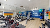 This fitness center is a comprehensively outfitted facility designed to support a full range of workout routines. The space is anchored by a bold blue accent wall displaying the motivational phrase "Fit. Fierce. Fabulous." in large white lettering, setting an energetic tone throughout. Floor-to-ceiling mirrors line one wall, visually expanding the space and allowing users to monitor their form during exercise.

The cardio zone features multiple treadmills, Echelon cycling bikes, and additional cardio equipment positioned on protective rubber mats over light hardwood-style flooring. A dual cable cross machine provides versatile strength training options, while an adjustable bench accommodates a variety of resistance exercises.

A dedicated accessories area houses a well-stocked ball rack with medicine balls, stability balls, and exercise balls of varying sizes, alongside a wall-mounted rack holding colorful yoga and foam rolling mats. A full dumbbell rack with tiered free weights rounds out the strength training offerings.

A vibrant color-block mural in shades of orange, yellow, and blue adds an artistic accent near the entrance, where a mounted flat-screen TV provides entertainment. Recessed and track lighting throughout ensures the space is well-lit, and an accessible restroom is conveniently located on-site. The overall ambiance is clean, modern, and motivating.
