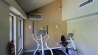 This fitness center offers a practical and functional workout environment contained within a single floor. The space is defined by warm, earthy tan-colored walls that create an inviting atmosphere, complemented by white trim and a dark rubberized flooring ideal for exercise equipment. The ceiling features an angled design that adds an architectural interest to the room, while wall-mounted sconce lighting provides ambient illumination throughout. A wall-mounted ventilation unit ensures proper air circulation during workouts. The cardio area is equipped with multiple exercise machines, including an elliptical trainer and a recumbent stationary bike, catering to a variety of cardiovascular fitness needs. A mirrored wall panel on one side of the room enhances the sense of space and allows users to monitor their form during exercise. A security camera is also installed, ensuring a safe and monitored environment. The overall ambiance is clean, focused, and purpose-driven, making this fitness center a convenient and efficient space for maintaining an active lifestyle.