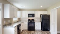 Located at 3340 Richlands Hwy in Jacksonville, NC, this two-story home offers a practical and comfortable layout designed for everyday living. The kitchen is a standout feature, outfitted with crisp white shaker-style cabinetry, granite-look countertops, and a full suite of black appliances including a gas range, over-the-range microwave, dishwasher, and refrigerator. Wood-look luxury vinyl plank flooring runs throughout the main living areas, complementing the warm, neutral-toned walls found throughout the home. The main level living area is open and airy, featuring a ceiling fan with light fixture, a staircase with white railings leading to the upper floor, and a window that allows natural light to fill the space. A back door in the kitchen area provides convenient exterior access. The upper level hosts carpeted bedrooms, each featuring ample natural light from windows and closet storage. One bedroom includes a ceiling fan, while another features a standard closet with built-in shelving. Bathrooms are cleanly finished with white fixtures and wood-look flooring. Throughout the home, white trim and neutral walls create a cohesive, move-in-ready aesthetic that is both versatile and inviting.