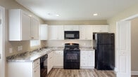 Located at 3340 Richlands Hwy in Jacksonville, NC, this two-story home offers a practical and comfortable layout designed for everyday living. The kitchen is a standout feature, outfitted with crisp white shaker-style cabinetry, granite-look countertops, and a full suite of black appliances including a gas range, over-the-range microwave, dishwasher, and refrigerator. Wood-look luxury vinyl plank flooring runs throughout the main living areas, complementing the warm, neutral-toned walls found throughout the home. The main level living area is open and airy, featuring a ceiling fan with light fixture, a staircase with white railings leading to the upper floor, and a window that allows natural light to fill the space. A back door in the kitchen area provides convenient exterior access. The upper level hosts carpeted bedrooms, each featuring ample natural light from windows and closet storage. One bedroom includes a ceiling fan, while another features a standard closet with built-in shelving. Bathrooms are cleanly finished with white fixtures and wood-look flooring. Throughout the home, white trim and neutral walls create a cohesive, move-in-ready aesthetic that is both versatile and inviting.