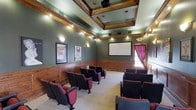 Located at 755 CL Tart Circle in Fayetteville, NC, this single-floor screening room delivers an immersive, theater-quality experience in an elegantly designed setting. The room is anchored by a large projection screen mounted on the front wall, complemented by a ceiling-mounted projector for high-quality viewing. Rows of classic red and black upholstered theater-style seating are arranged to provide unobstructed sightlines throughout the space. The interior design showcases rich wood wainscoting that lines the lower half of the walls, paired with deep sage green upper walls that create a warm, intimate ambiance. A striking coffered ceiling with dark wood beams adds architectural depth and character overhead, while recessed and accent lighting is thoughtfully positioned to illuminate the space without detracting from the viewing experience. Vintage-style movie posters adorn the walls, reinforcing the classic cinema aesthetic. A draped window with rich burgundy curtains on the side wall allows for natural light control. The carpeted floor and carefully curated design elements work together to provide both comfort and excellent acoustics, making this a standout venue for private screenings and entertainment.