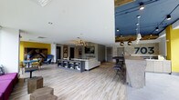 This single-floor space showcases a contemporary open-concept design that seamlessly blends multiple functional zones within one expansive area. Light wood-style flooring runs throughout, contributing to a bright and airy atmosphere complemented by white ceilings with recessed lighting. A central lounge area features a low-profile white sofa paired with a built-in media console offering open shelving for books and decorative items, anchored by a wall-mounted flat-screen TV set against a bold geometric patterned accent wall. A statement chandelier with warm Edison-style bulbs adds an elegant focal point overhead. To one side, a reception or bar-height counter finished in a textured aggregate surface is paired with sleek black barstools and pendant lighting, creating a social gathering spot. A bold yellow accent wall and a striking blue lounge chair add pops of color, enhancing the eclectic, modern aesthetic. Reclaimed wood ceiling panels and dark navy blue architectural details provide contrast and visual depth throughout. The open layout encourages natural circulation between zones, making the space feel both dynamic and inviting for a variety of uses.