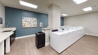 Located at 225 W Third St in Long Beach, California, this shared laundry facility is a thoughtfully designed, single-floor space built for convenience and comfort. The room is finished with light wood-look flooring and a drop ceiling fitted with recessed panel lighting that ensures bright, even illumination throughout.

The facility is divided into two distinct machine zones. One section houses a row of front-loading washing machines and dryers lined neatly along the wall, while a second area features top-loading washers, each equipped with digital control panels for ease of use. The machines are arranged for efficient workflow, with ample space to move between them.

A generous built-in folding counter runs along one wall, providing residents with a dedicated surface for sorting and folding laundry. A centrally located partition column adds structural definition to the space without disrupting its open feel. Accent walls painted in a calming steel blue add a touch of color and visual interest, complemented by decorative artwork including coastal-themed and abstract prints.

Informational bulletin boards and posted notices keep residents informed of building updates and facility guidelines. A utility closet with glass-paneled doors offers additional storage. Trash receptacles are conveniently placed throughout. The overall design is clean, modern, and functional, reflecting a well-maintained shared amenity space.